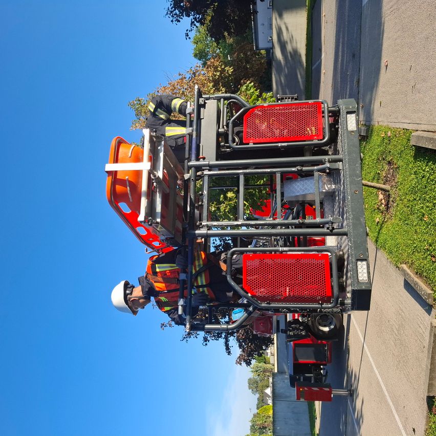 Zwei Feuerwehrleute arbeiten auf einem Fahrzeug an einer Straße mit grünem Gras und Bäumen. Das Fahrzeug hat rote und schwarze Teile und einen Sicherheitskäfig.