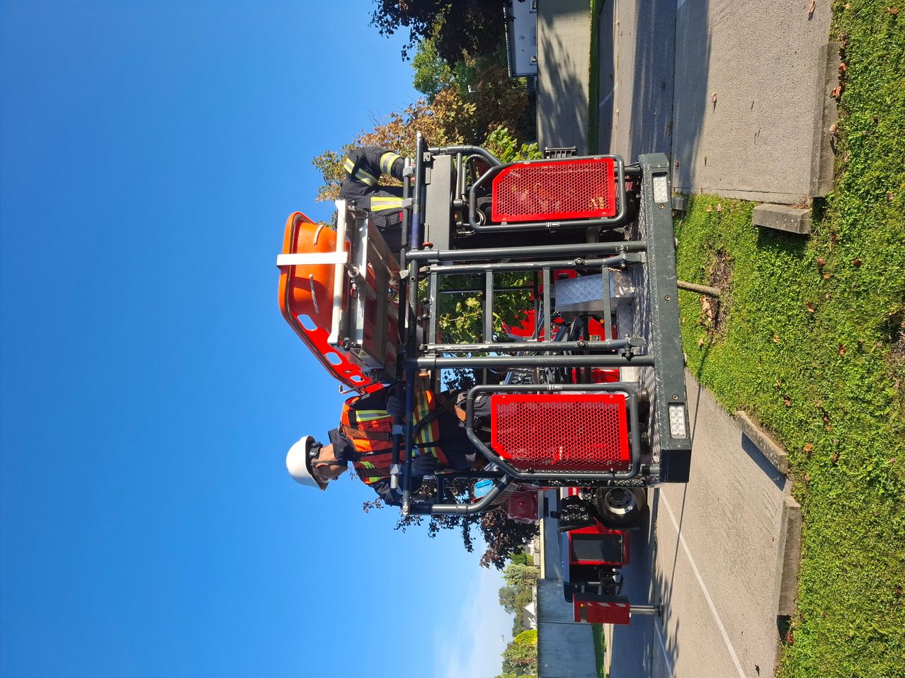 Zwei Feuerwehrleute arbeiten auf einem Fahrzeug an einer Straße mit grünem Gras und Bäumen. Das Fahrzeug hat rote und schwarze Teile und einen Sicherheitskäfig.