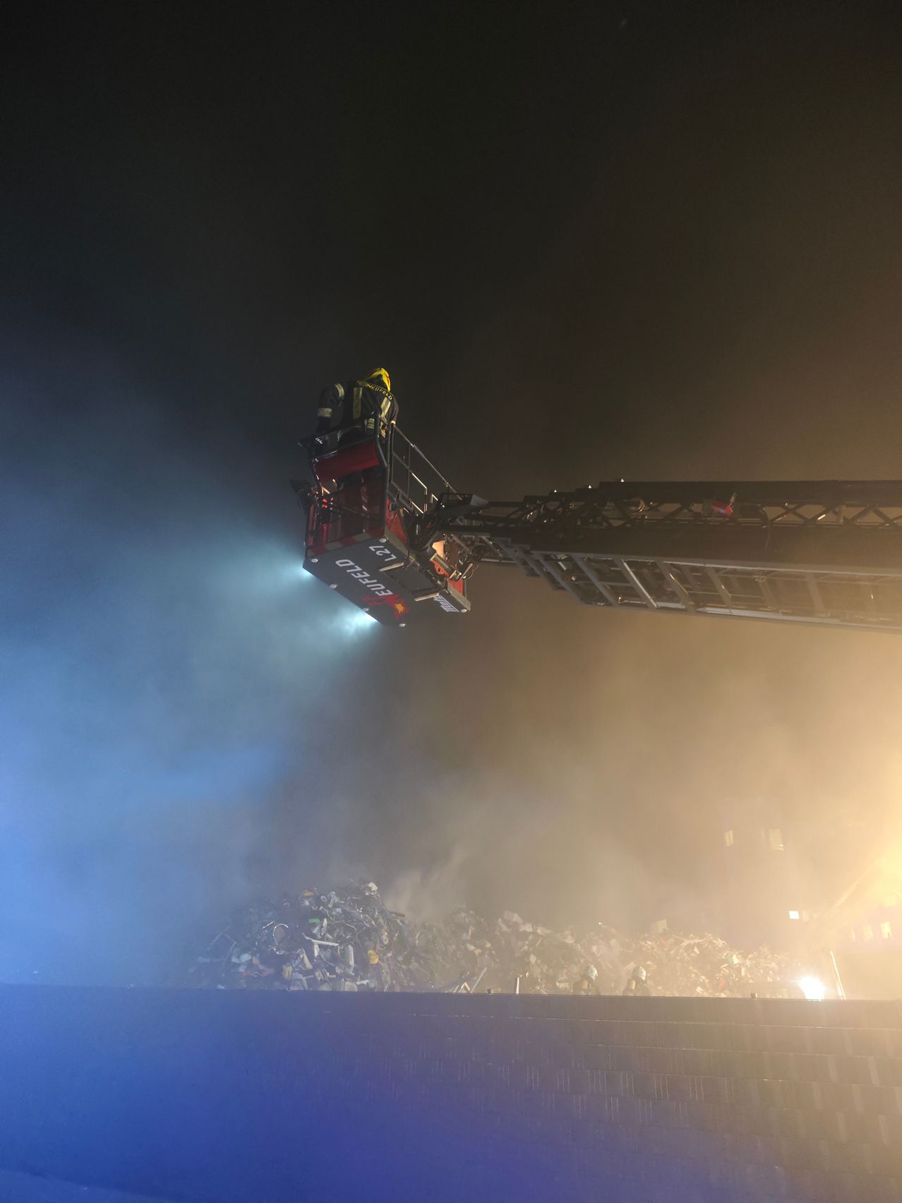 Feuerwehrleute auf einer Drehleiter schweben bei Nacht über einem brennenden Schrottplatz. Die Drehleiter gibt ein starkes Licht ab.