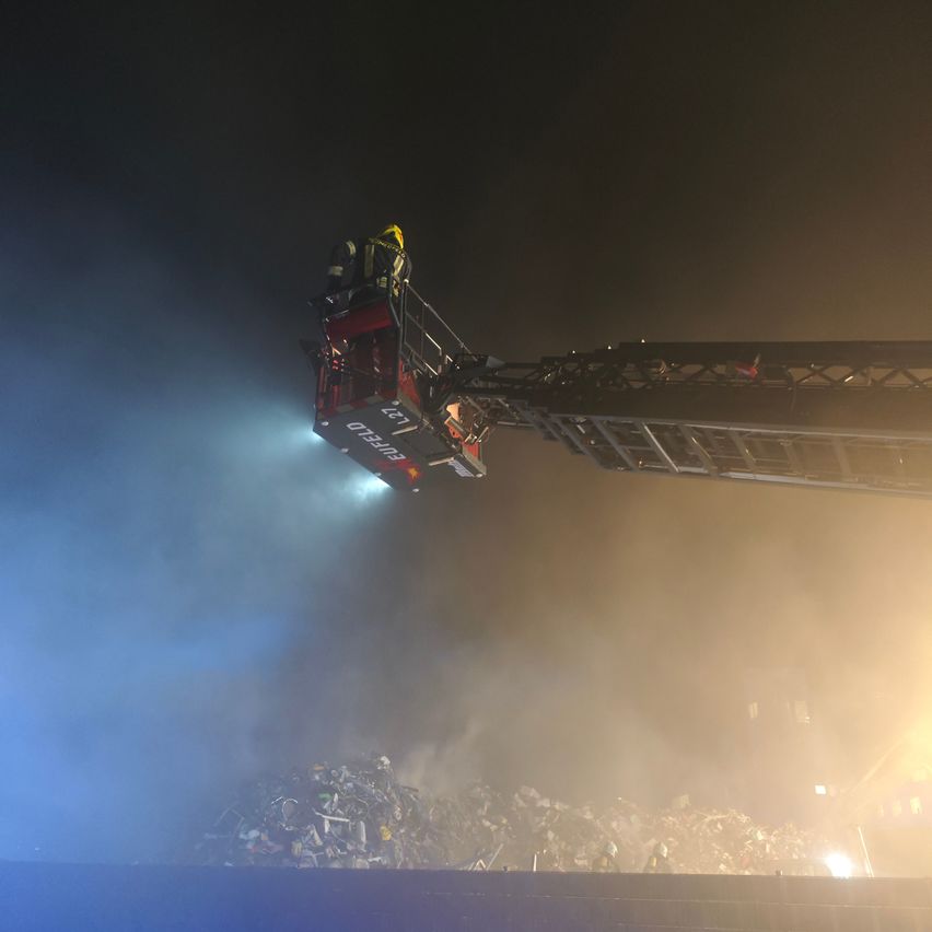 Feuerwehrleute auf einer Drehleiter schweben bei Nacht über einem brennenden Schrottplatz. Die Drehleiter gibt ein starkes Licht ab.