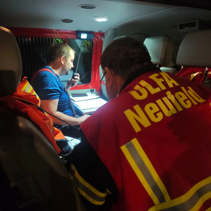 Zwei Feuerwehrleute in einem Fahrzeug, einer spricht in ein Funkgerät, während der andere ein Dokument betrachtet. Die Person im roten Jackett trägt den Namen Neufeld.