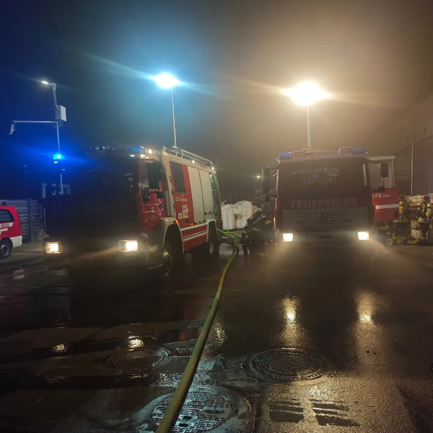 Zwei Feuerwehrwagen sind nachts auf einer nassen Straße geparkt, wobei Feuerwehrleute einen Schlauch an einen der Wagen anschließen.