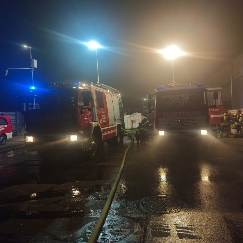 Zwei Feuerwehrwagen sind nachts auf einer nassen Straße geparkt, wobei Feuerwehrleute einen Schlauch an einen der Wagen anschließen.