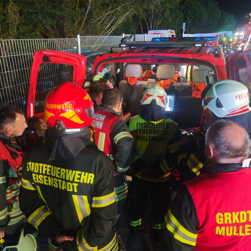 Feuerwehrleute in roten und gelben Uniformen stehen um ein rotes Fahrzeug mit offener Tür. Eine Person mit Helm ist im Fahrzeug. Ein Mann mit rotem Weste steht rechts.
