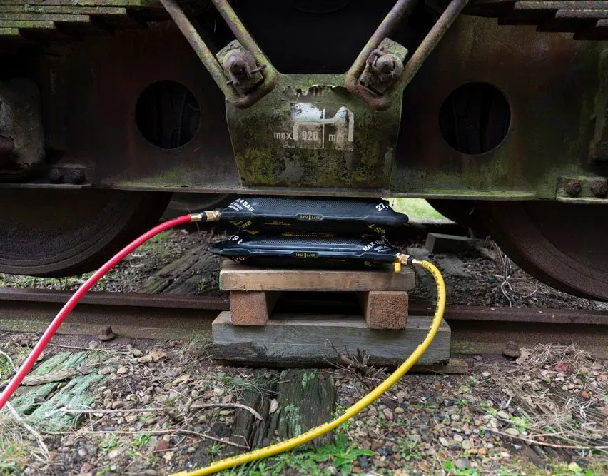 Zwei schwarze Boxen mit angeschlossenen Rohren stehen auf Holzblöcken, befestigt an einem rostigen Zug auf einer Eisenbahn.