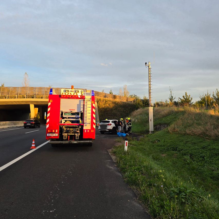 Ein Feuerwehrauto ist am Straßenrand geparkt, in der Nähe ein weißes Auto. Einige Leute stehen in der Nähe des Autos. Ein Verkehrskegel steht am Straßenrand.