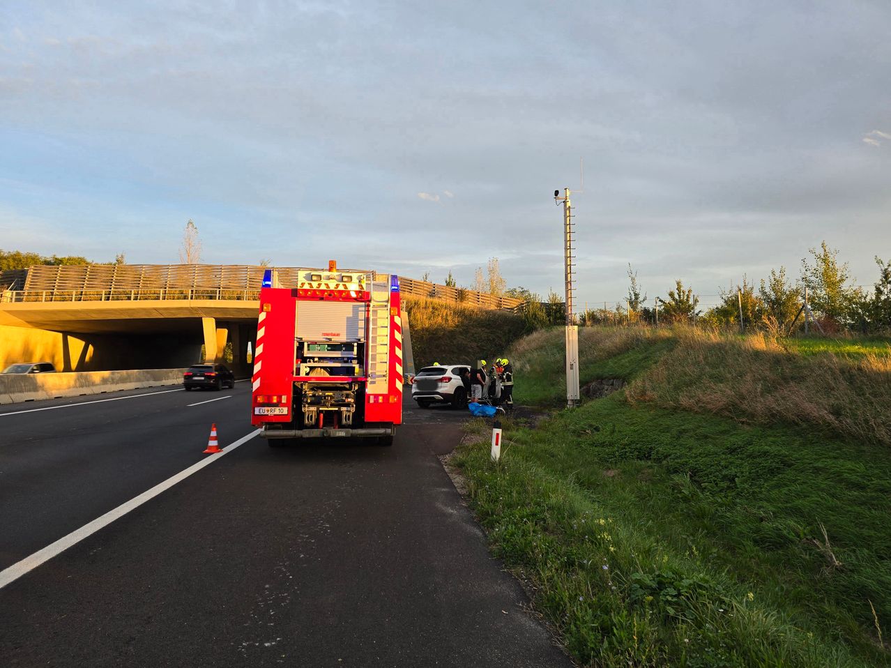 Ein Feuerwehrauto ist am Straßenrand geparkt, in der Nähe ein weißes Auto. Einige Leute stehen in der Nähe des Autos. Ein Verkehrskegel steht am Straßenrand.