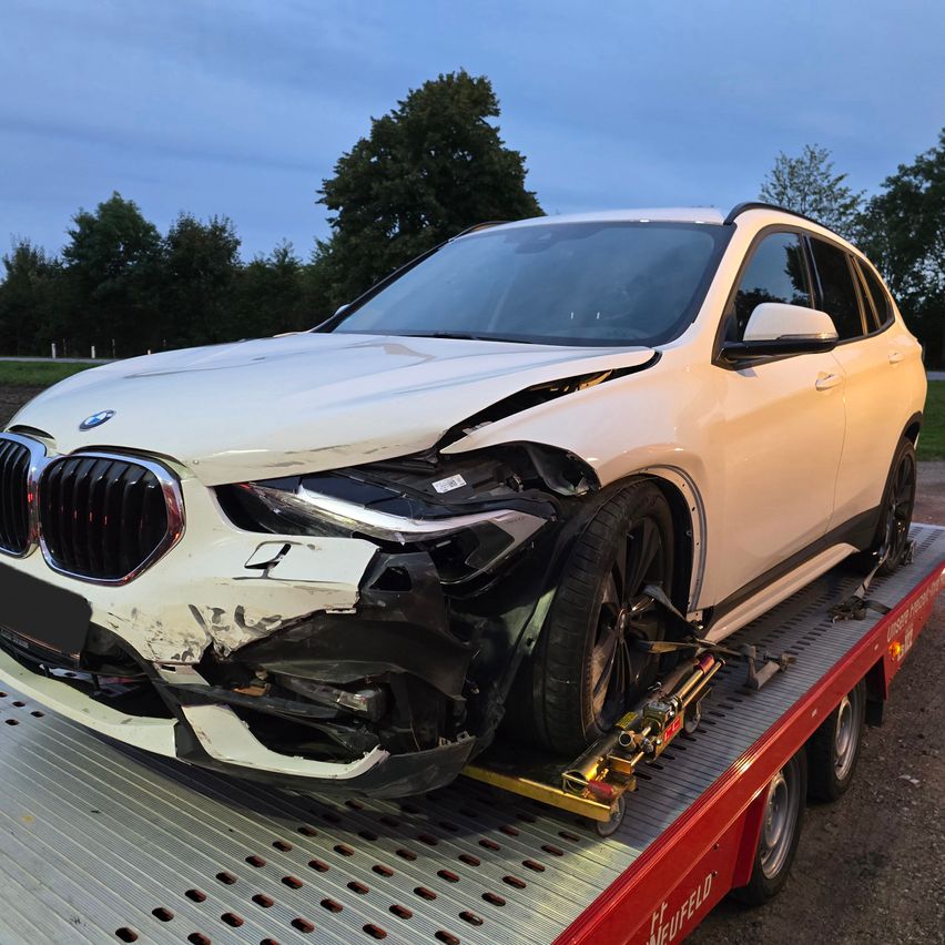 Ein weißer BMW X1 SUV steht auf einem roten Abschleppwagen mit beschädigter Front. Der SUV ist mit einem Abschlepphaken gehoben und hat einen beschädigten Stoßfänger, Scheinwerfer und Kühlergrill.