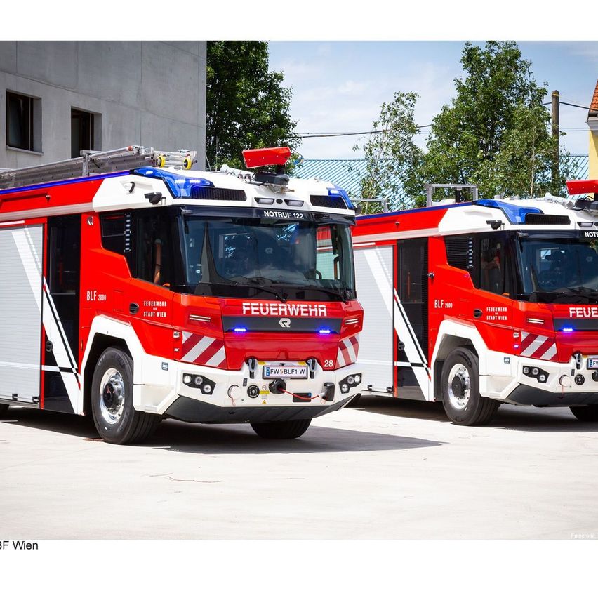 Zwei rote Feuerwehrfahrzeuge sind vor einem Gebäude geparkt. Sie sind mit dem Wort 'Feuerwehr' beschriftet, und eines von ihnen hat das Kennzeichen 'FW/BLF1'. Im Hintergrund befinden sich Bäume und ein Haus.