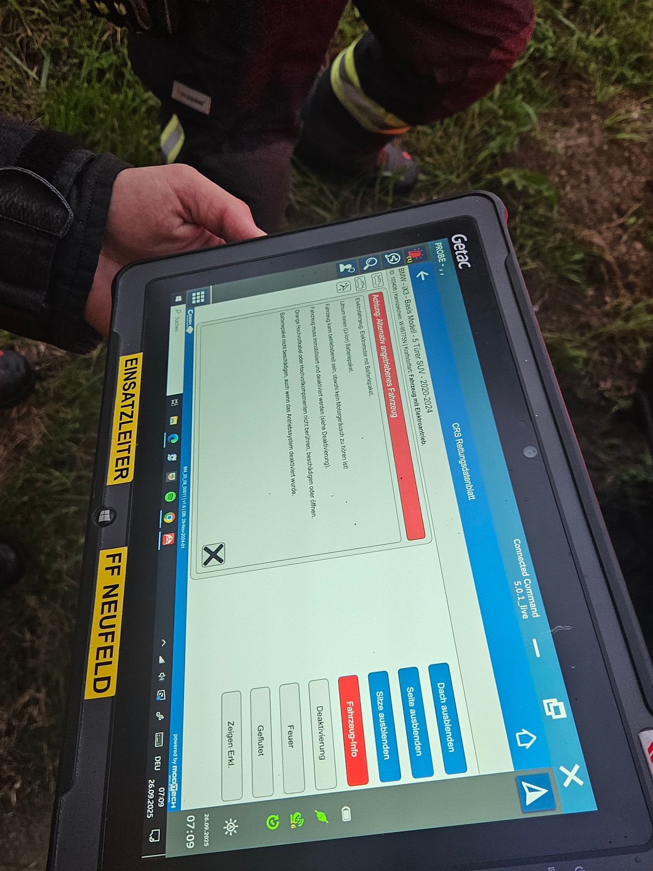 Ein Tablet-Gerät zeigt eine Seite mit einem roten Kreuz und dem Text 'EINSATZLETTER' an der Seite. Es wird von jemandem mit einem grasigen Hintergrund gehalten.