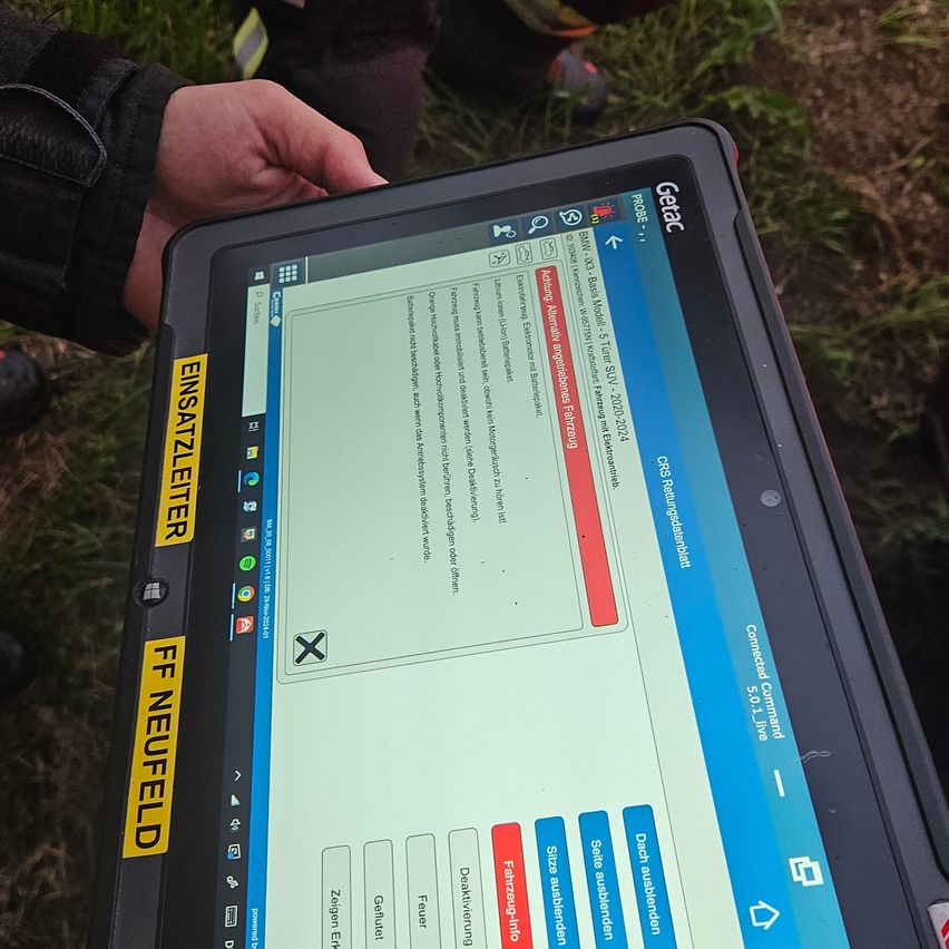 Ein Tablet-Gerät zeigt eine Seite mit einem roten Kreuz und dem Text 'EINSATZLETTER' an der Seite. Es wird von jemandem mit einem grasigen Hintergrund gehalten.