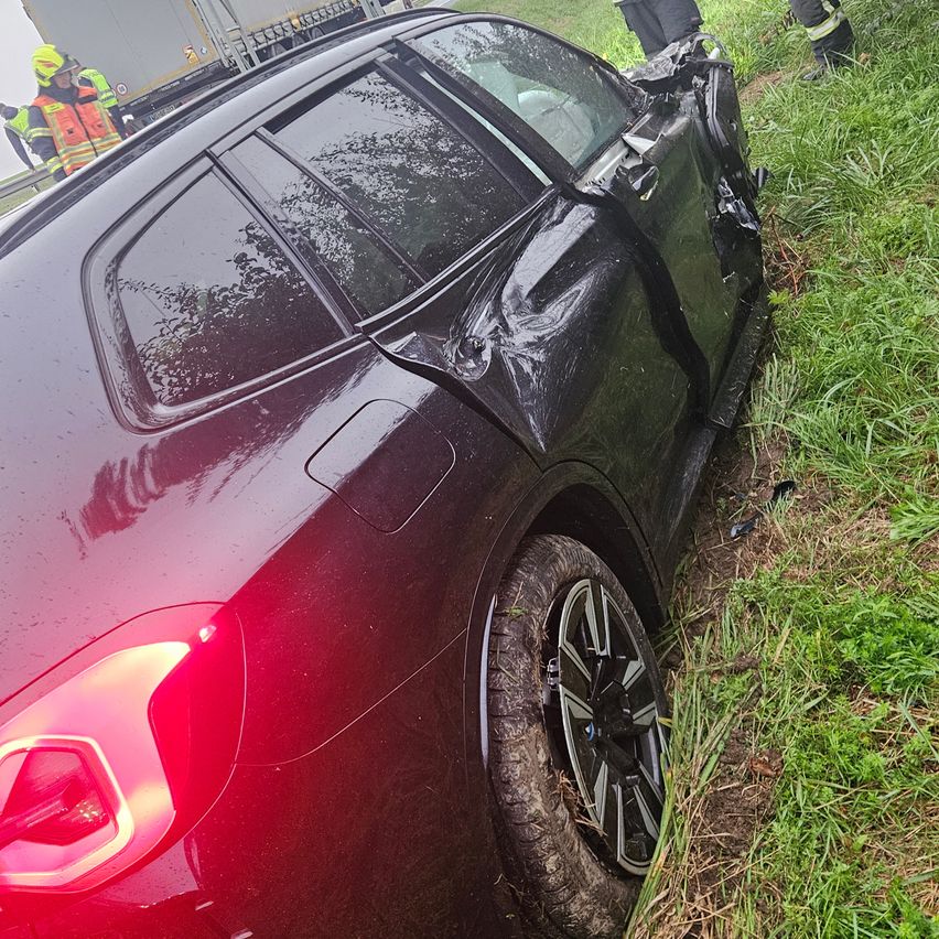 Ein kastanienbraunes Auto ist am Straßenrand beschädigt, wobei das Vorderrad abgekoppelt ist. Ein LKW steht dahinter, und zwei Feuerwehrleute sind in der Nähe.