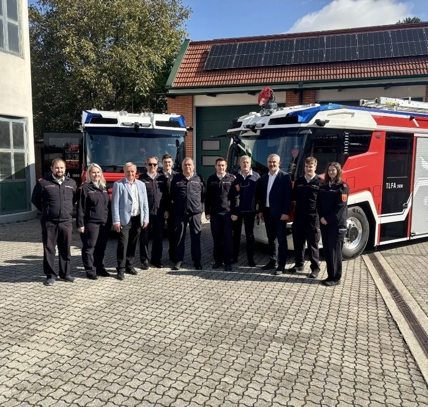 Eine Gruppe von Feuerwehrleuten und Beamten steht vor einem Feuerwehrauto mit Solarpanelen auf dem Dach der Feuerwache.
