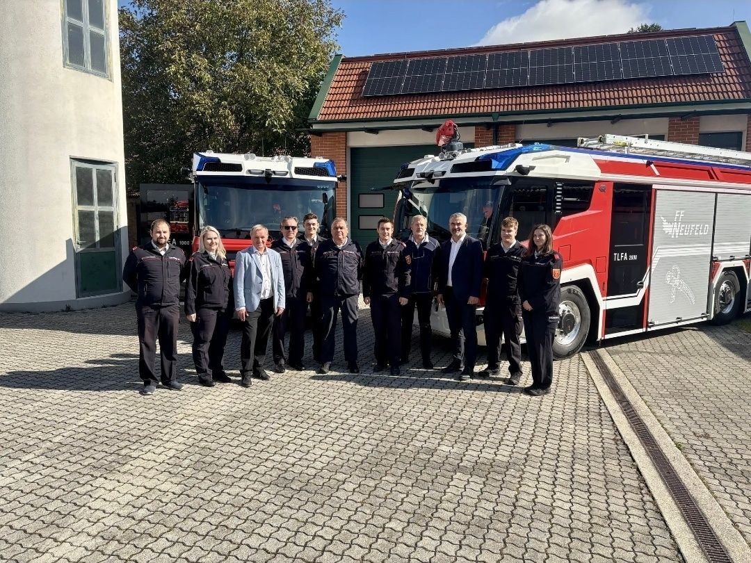 Eine Gruppe von Feuerwehrleuten und Beamten steht vor einem Feuerwehrauto mit Solarpanelen auf dem Dach der Feuerwache.