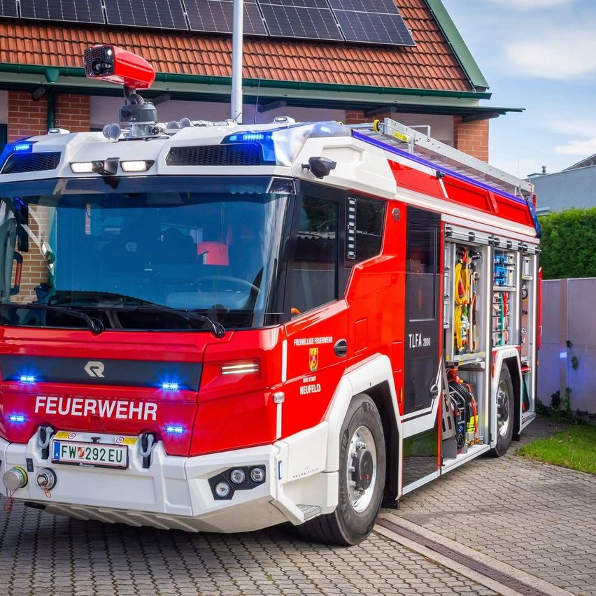 Ein rot-weißer Feuerwehrwagen mit blauen Lichtern steht vor einem Backsteingebäude mit Solarpanelen auf dem Dach.