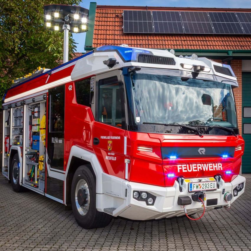 Ein roter Feuerwehrwagen mit der Aufschrift 'FEUERWEHR' steht vor einem Gebäude mit Solarmodulen auf dem Dach.