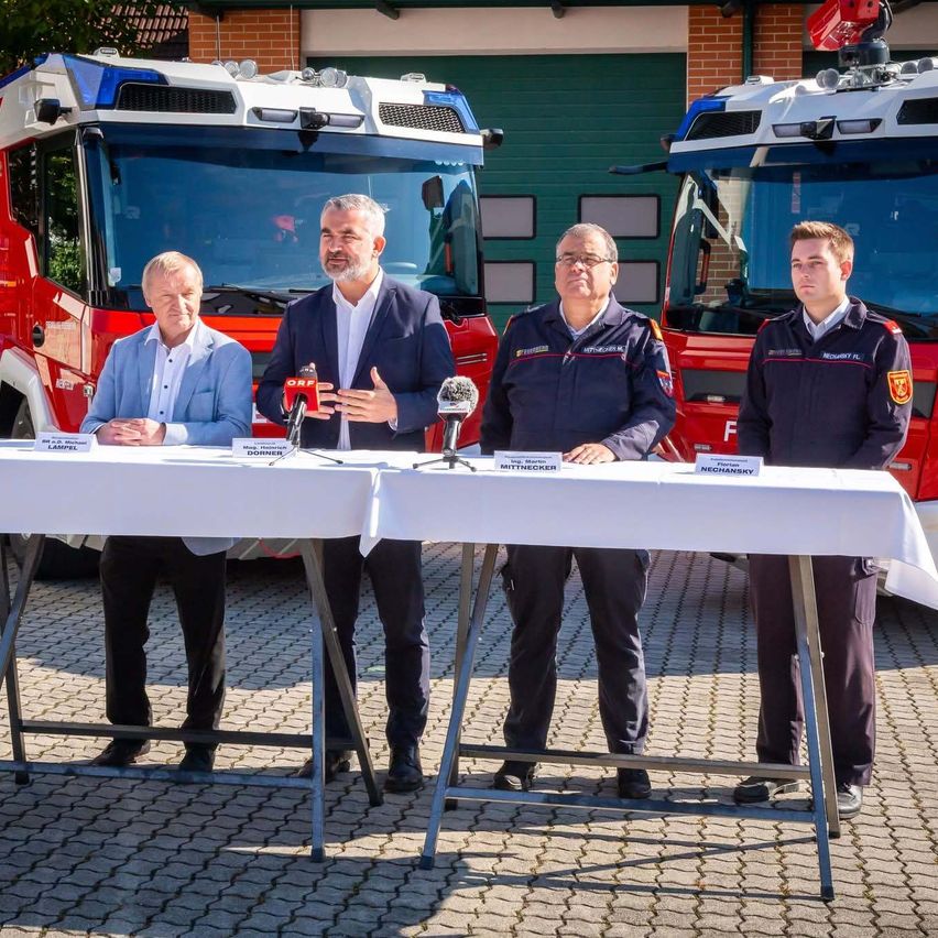Vier Männer stehen vor einem Tisch vor zwei Feuerwehrautos. Sie scheinen eine Pressekonferenz abzuhalten.