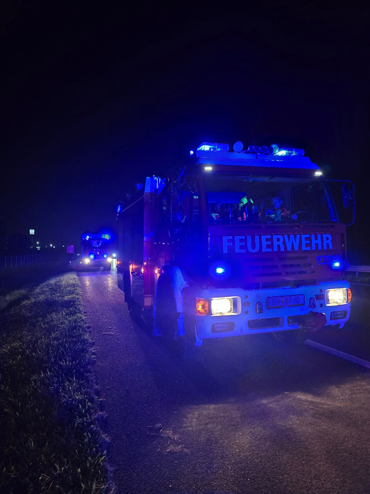 Ein Feuerwehrfahrzeug fährt nachts mit eingeschalteten blauen Lichtern auf einer Straße. In der Ferne ist ein weiteres Fahrzeug mit eingeschalteten Lichtern sichtbar.