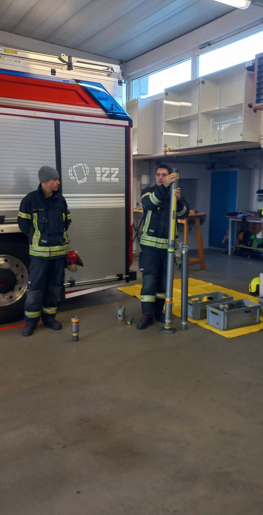 Zwei Feuerwehrleute in Uniform stehen in einer Garage. Einer hält einen Stab, der andere steht neben einem Feuerwehrwagen. Verschiedene Ausrüstungsgegenstände und Lagerboxen sind um sie herum.