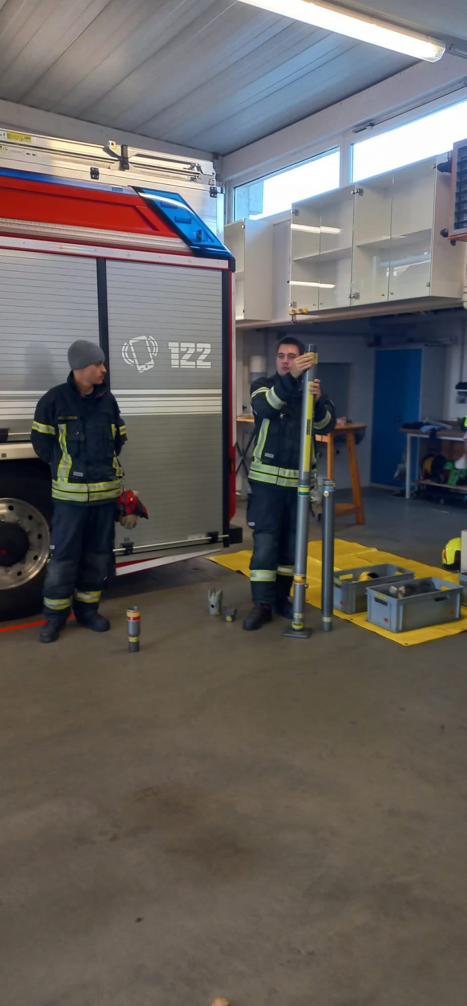 Zwei Feuerwehrleute in Uniform stehen in einer Garage. Einer hält einen Stab, der andere steht neben einem Feuerwehrwagen. Verschiedene Ausrüstungsgegenstände und Lagerboxen sind um sie herum.