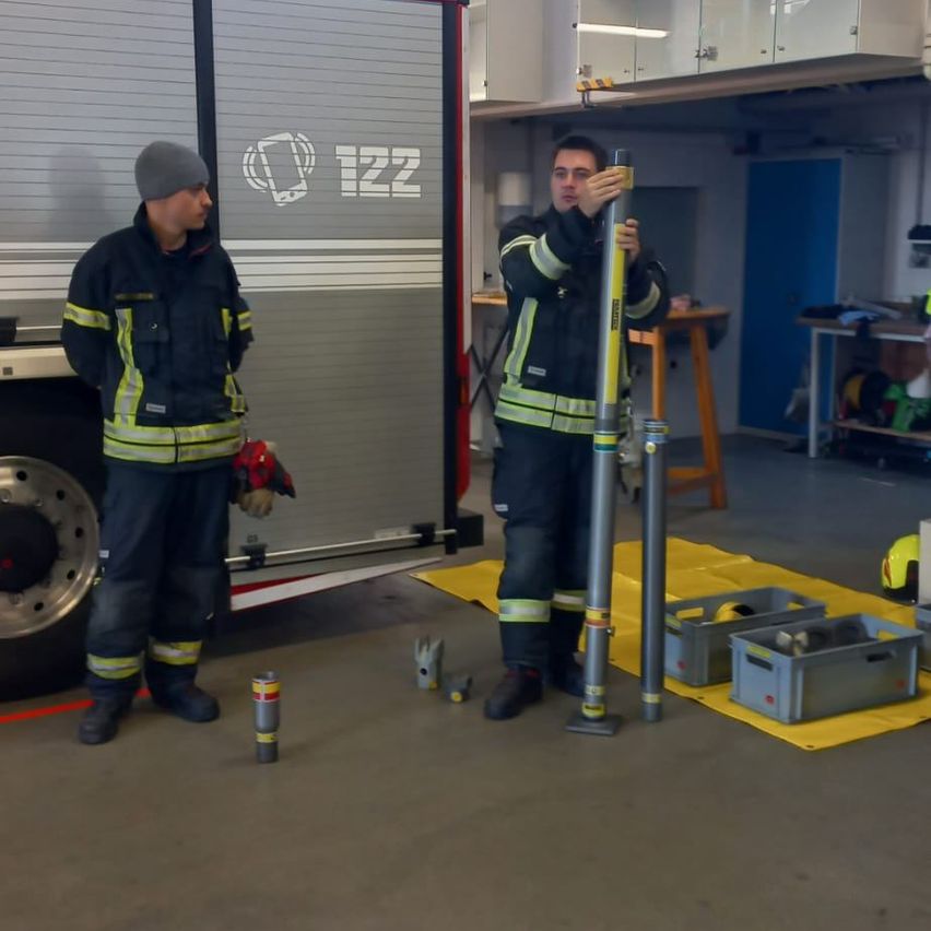 Zwei Feuerwehrleute in Uniform stehen in einer Garage. Einer hält einen Stab, der andere steht neben einem Feuerwehrwagen. Verschiedene Ausrüstungsgegenstände und Lagerboxen sind um sie herum.