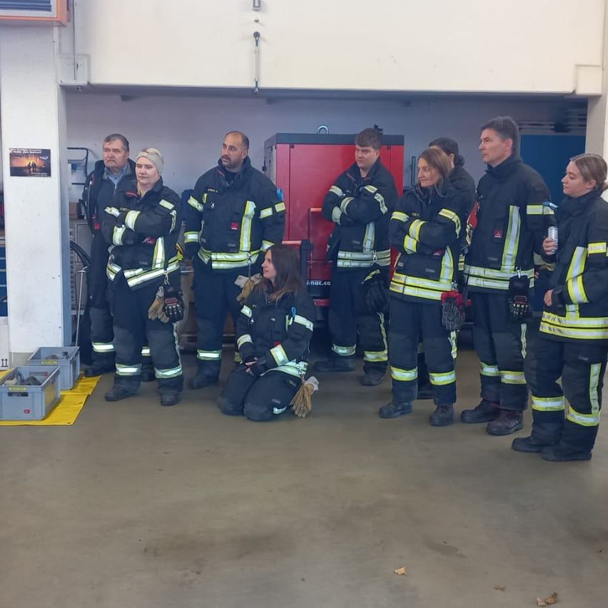 Eine Gruppe von Feuerwehrleuten, sowohl Männer als auch Frauen, steht in einer Garage, wobei eine Frau kniet, umgeben von verschiedenen Werkzeugen und Ausrüstungsgegenständen.