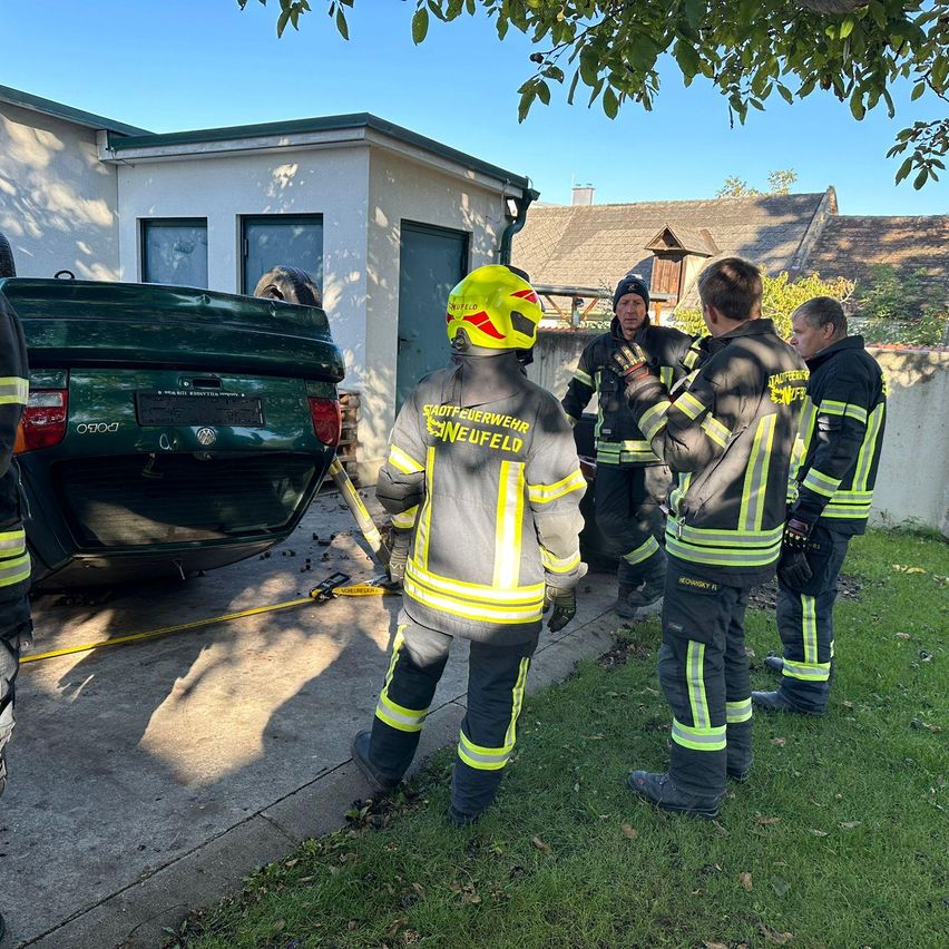 Ein Feuerwehrteam steht neben einem grünen Auto mit beschädigtem Heck vor einem Haus.