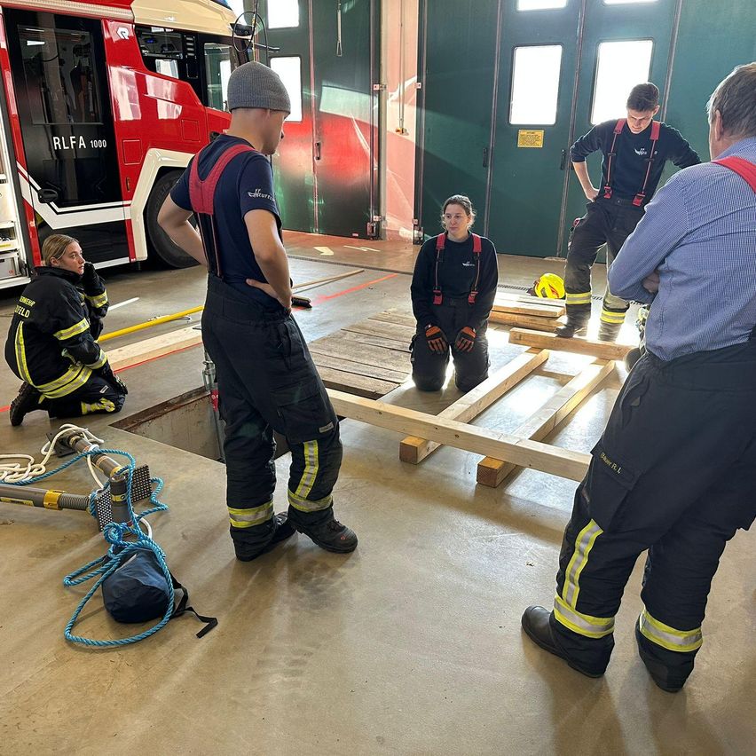 Eine Gruppe von Feuerwehrleuten in voller Ausrüstung übt in einer Feuerwache. Einer kauert, ein anderer steht mit den Händen in den Hüften, und zwei weitere beobachten. Ein Feuerwehrwagen ist in der Nähe geparkt.