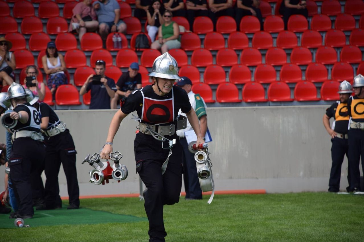 Ein Feuerwehrmann in Uniform, mit Ausrüstung, läuft über ein Feld, während Zuschauer im Hintergrund zu sehen sind.