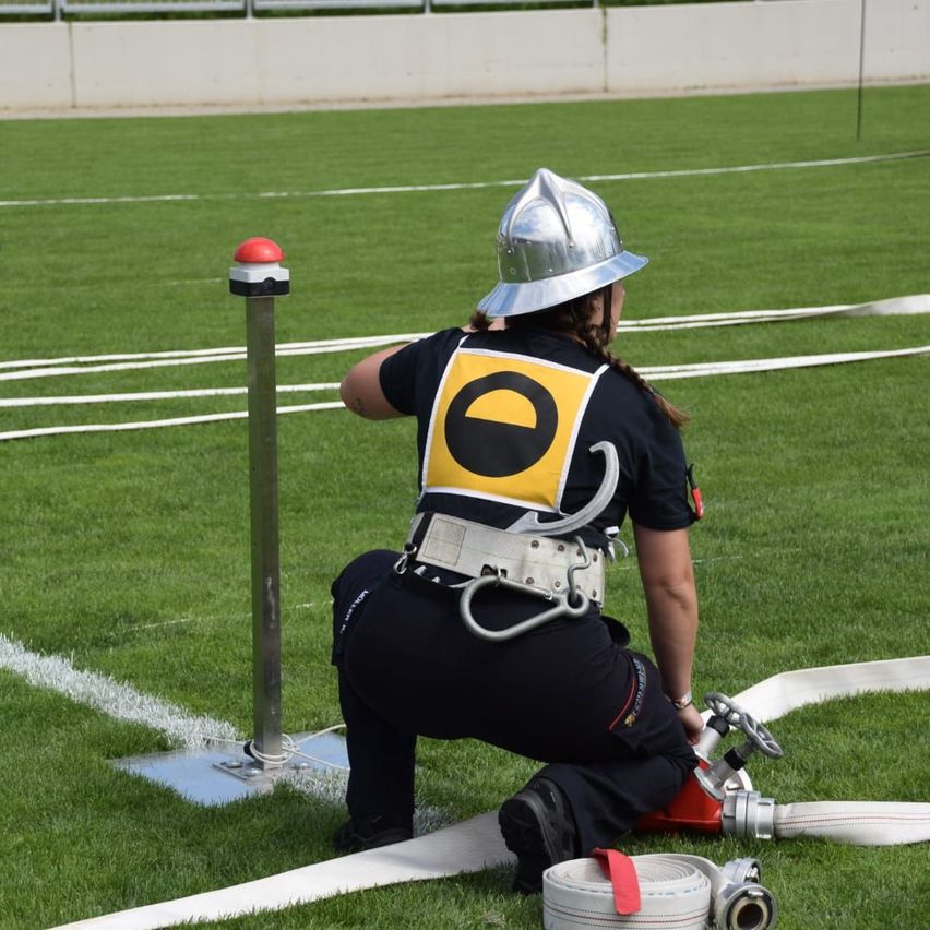 Ein Feuerwehrmann, der einen silbernen Helm trägt, hockt auf einem grünen Feld und repariert einen Feuerhydranten mit einem Schlauch in der Nähe.