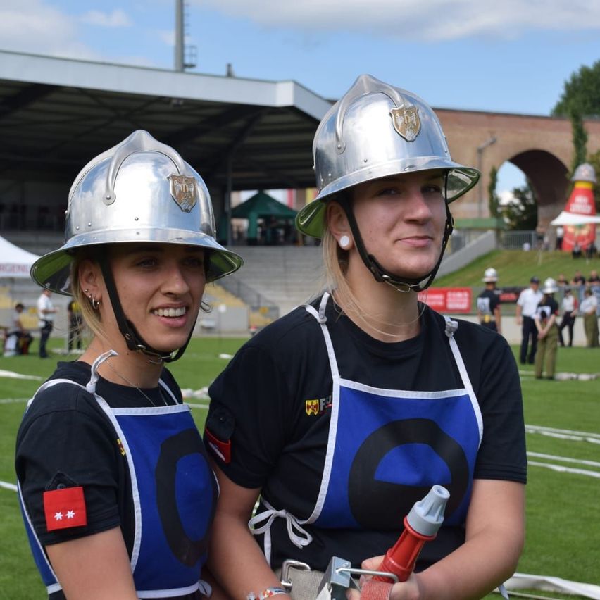 Zwei Frauen in Feuerwehruniformen und Helmen posieren für ein Foto auf einem Feld. Sie haben Abzeichen auf ihren Schürzen. Hinter ihnen steht ein Stadion und stehende Personen.