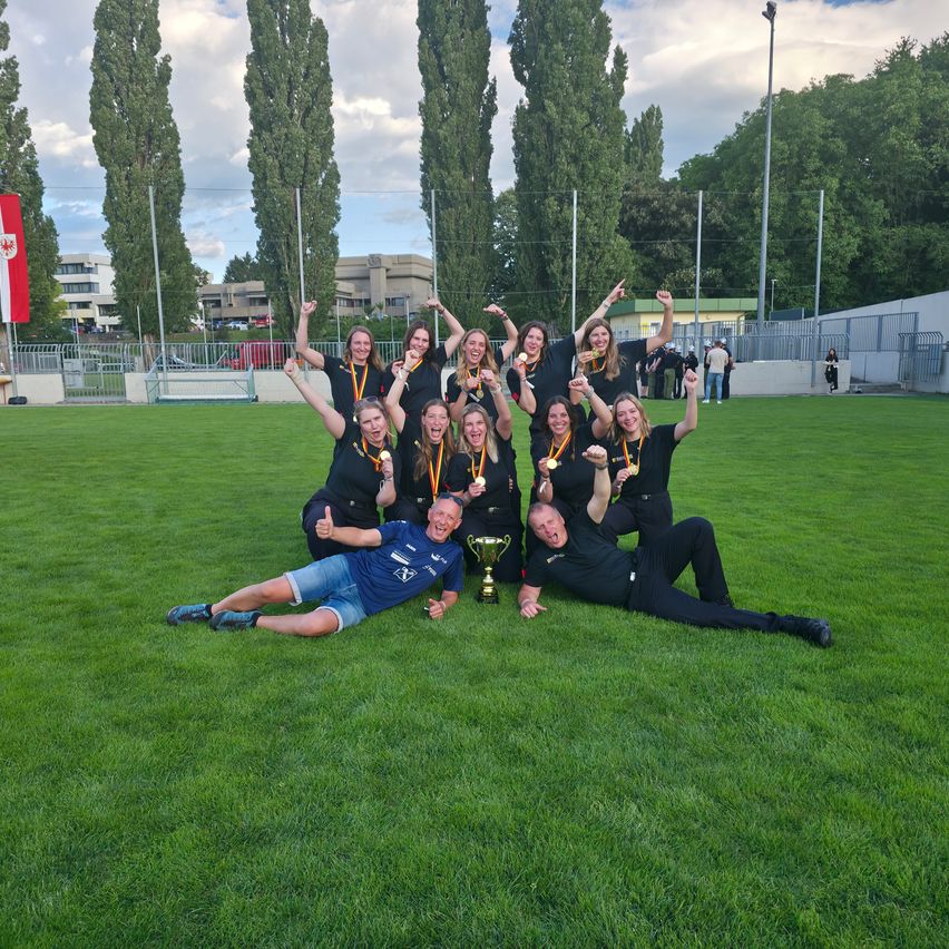 Eine Gruppe von Athleten feiert auf einem Feld mit einem Pokal vor ihnen, mit Gebäuden und Bäumen im Hintergrund.