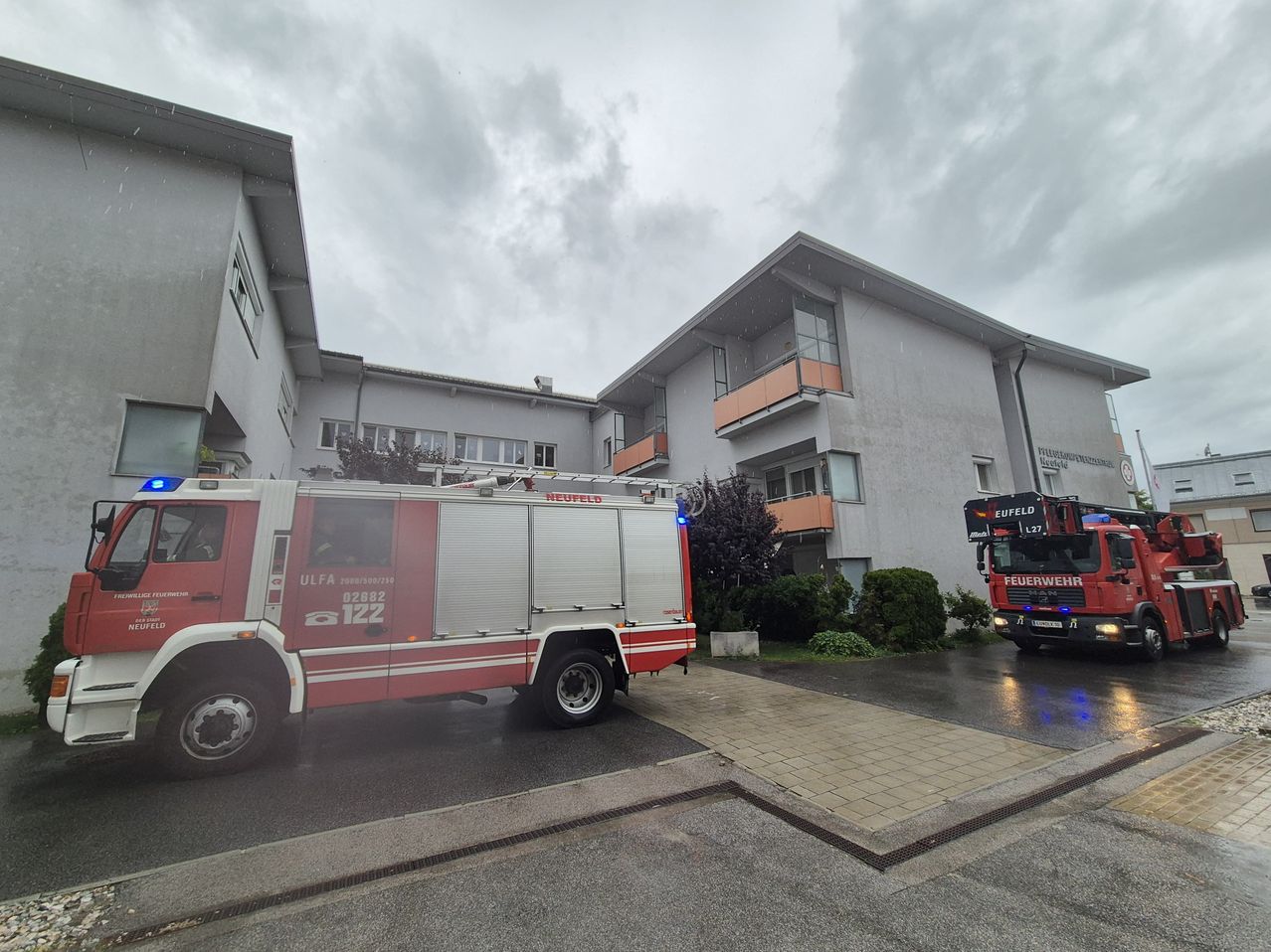 Zwei rote Feuerwehrfahrzeuge sind vor einem Gebäude mit Balkonen und einem bewölkten Himmel geparkt.