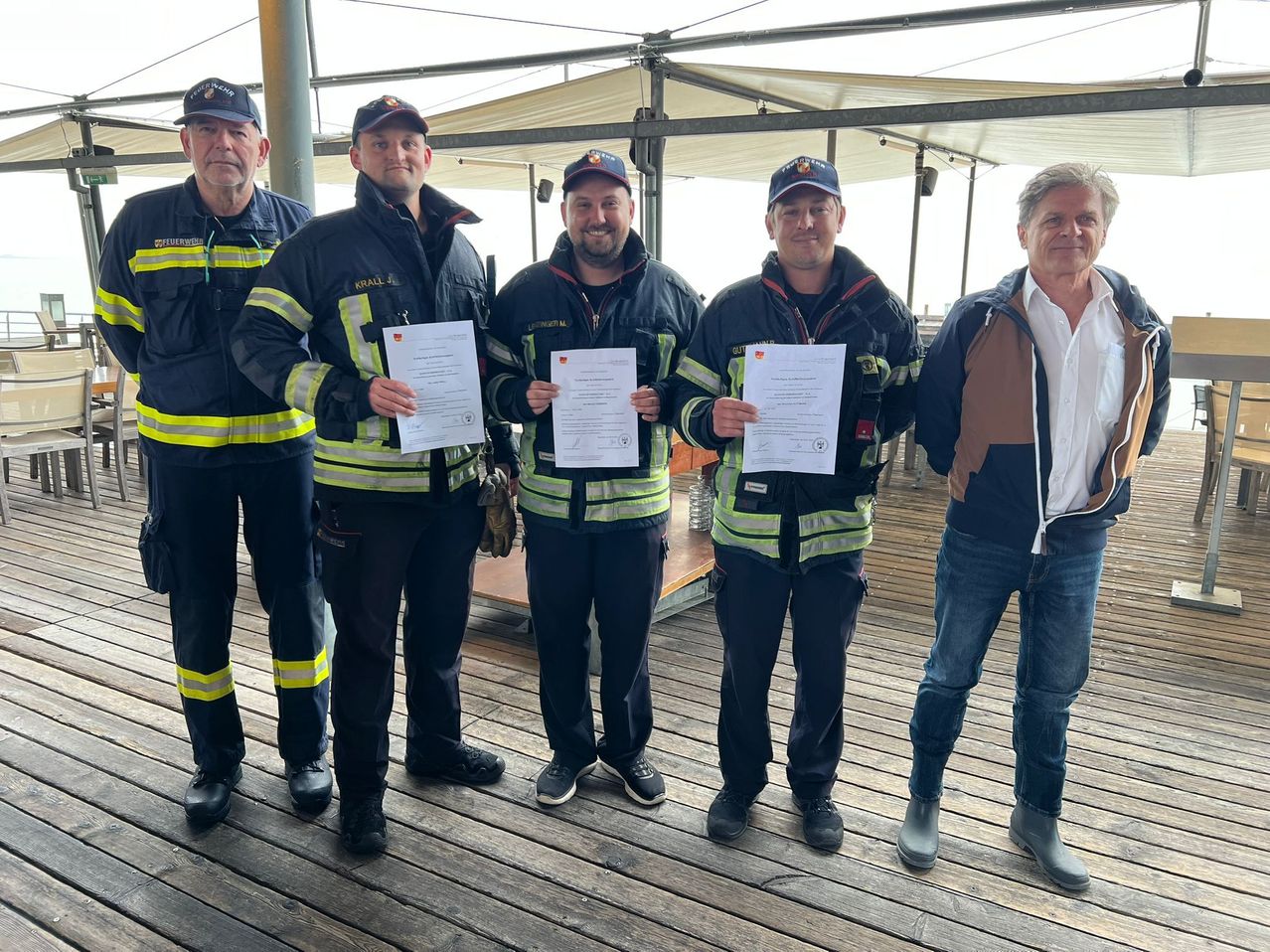 Fünf Männer in Uniformen stehen auf einer Holzplattform. Sie halten Urkunden in den Händen, einer in einem weißen Hemd. Hinter ihnen ist ein Zelt.