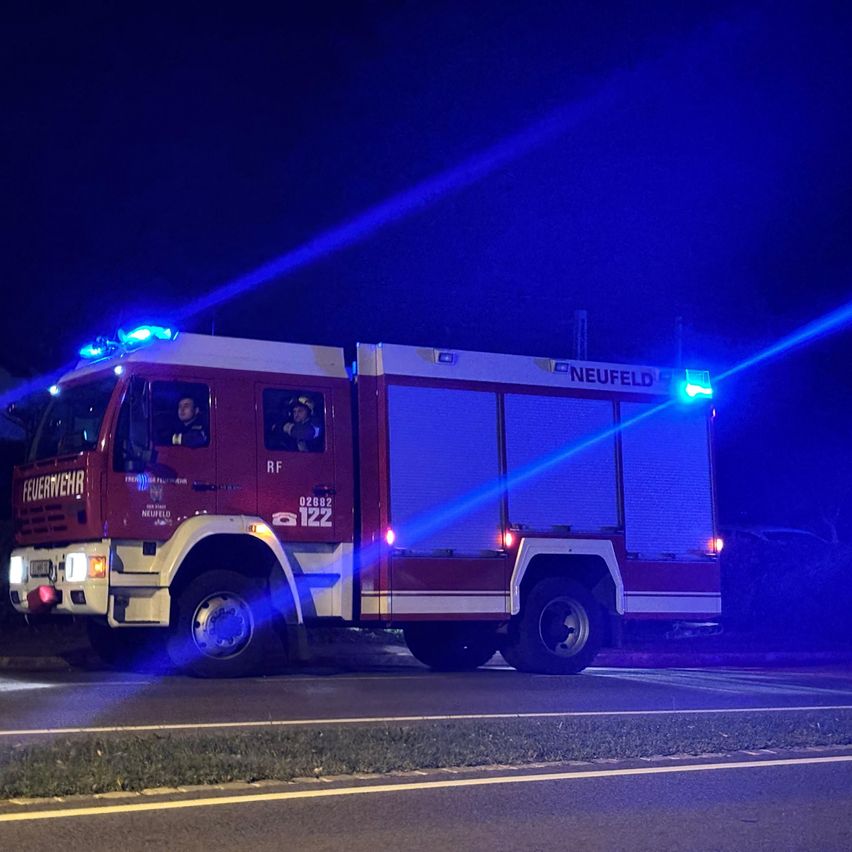 Ein rotes Notfallfahrzeug aus Neufeld mit blauen Lichtern steht nachts auf der Straße. Zwei Personen sind im Fahrzeug.