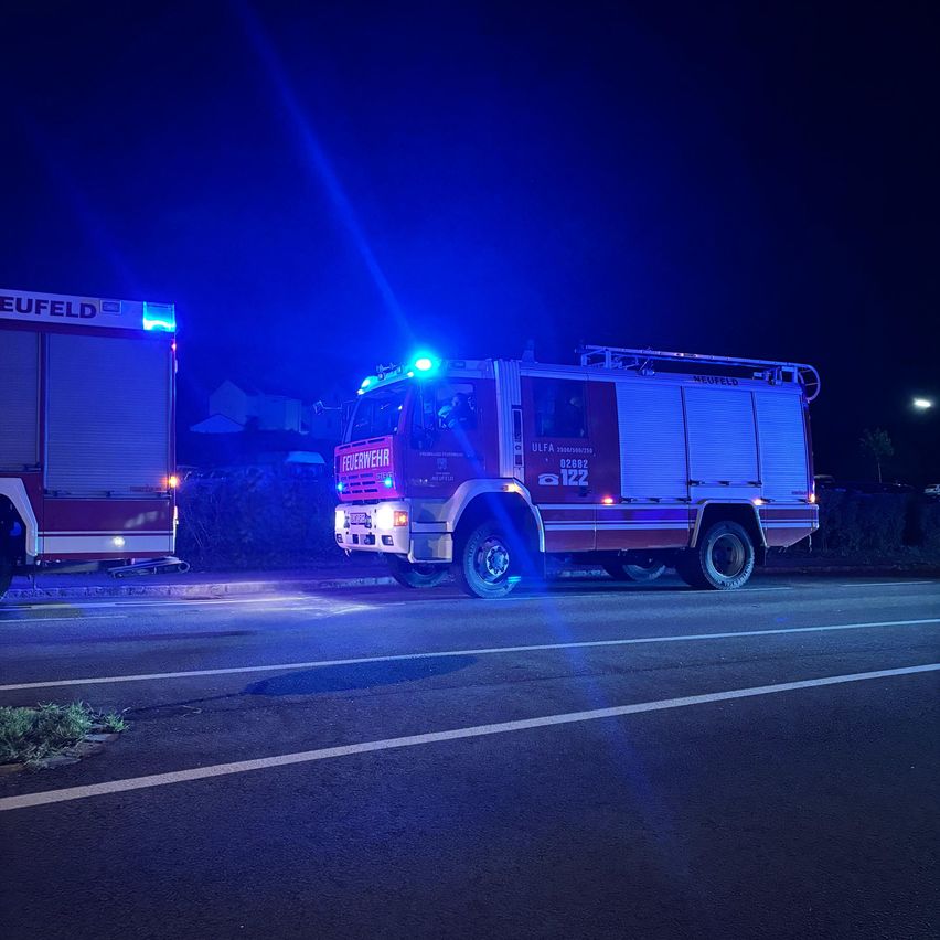 Zwei Feuerwehrfahrzeuge sind am Rande einer dunklen Straße in der Nacht geparkt. Das vordere hat seine Lichter eingeschaltet, und das andere ist teilweise auf der linken Seite sichtbar.