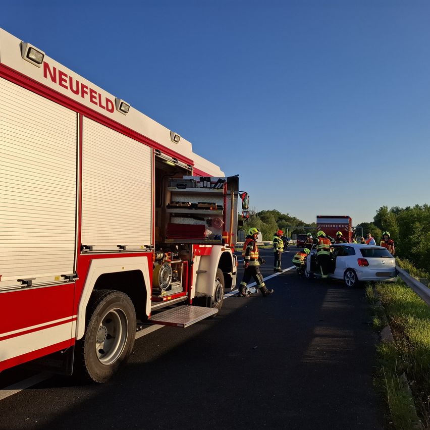 Ein Feuerwehrfahrzeug mit der Aufschrift Neufeld steht am Straßenrand, während Rettungskräfte sich um einen weißen Unfallwagen kümmern.
