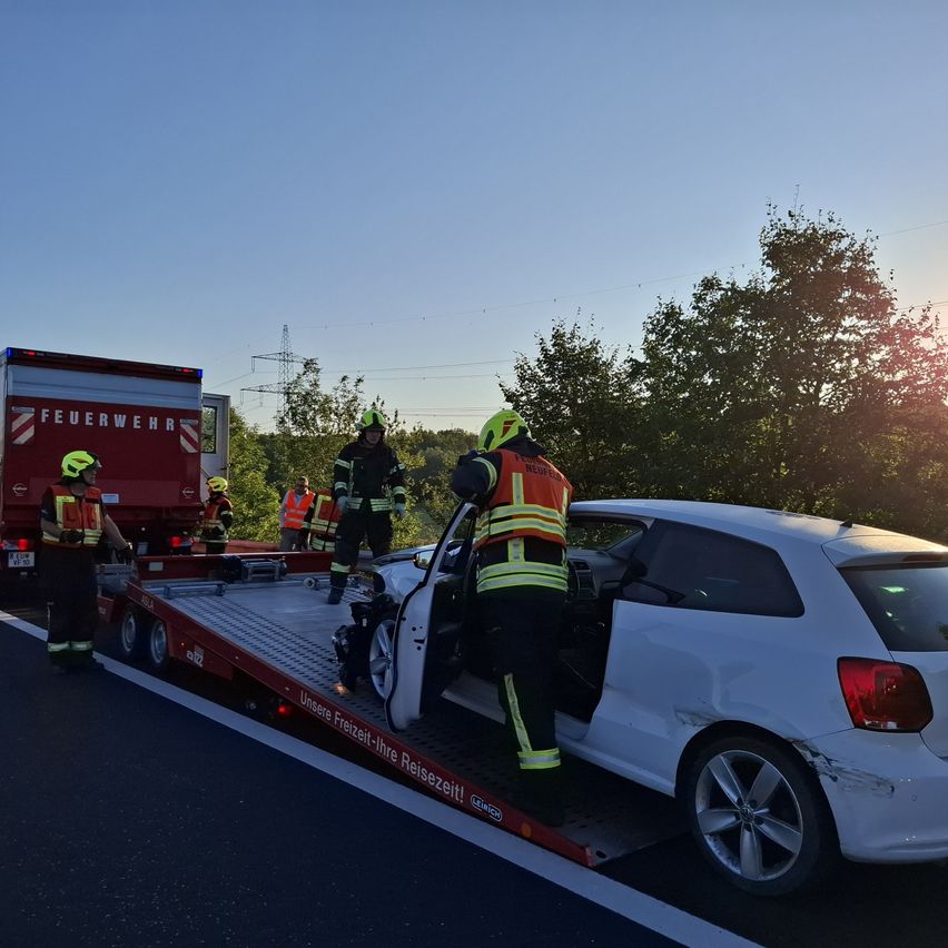 Feuerwehrleute entfernen ein beschädigtes Auto von der Straße. Ein roter Abschleppwagen zieht das Fahrzeug. Ein Feuerwehrmann steht im Auto, während andere am Straßenrand stehen.