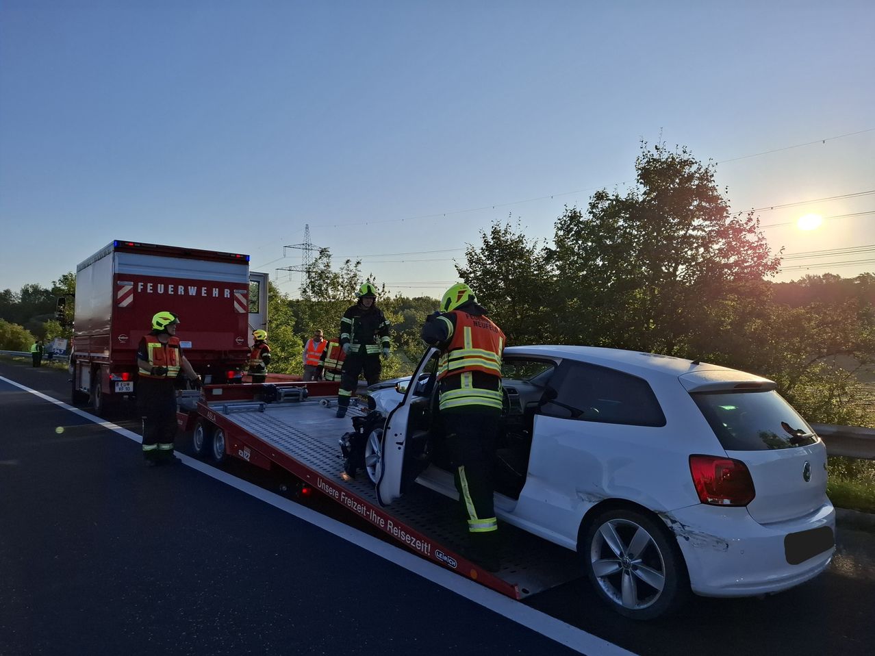 Feuerwehrleute entfernen ein beschädigtes Auto von der Straße. Ein roter Abschleppwagen zieht das Fahrzeug. Ein Feuerwehrmann steht im Auto, während andere am Straßenrand stehen.