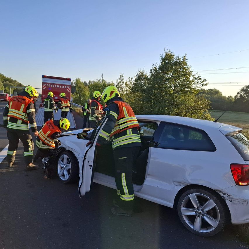 Rettungskräfte kümmern sich um ein beschädigtes weißes Auto am Straßenrand. Ein Feuerwehrwagen steht hinter ihnen.