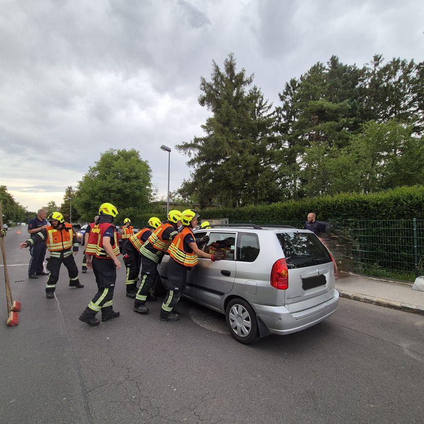 Feuerwehrleute in gelben Helmen schieben ein silbernes Auto an einem bewölkten Tag. Bäume und Büsche sind in der Umgebung, mit einem Zaun im Hintergrund.