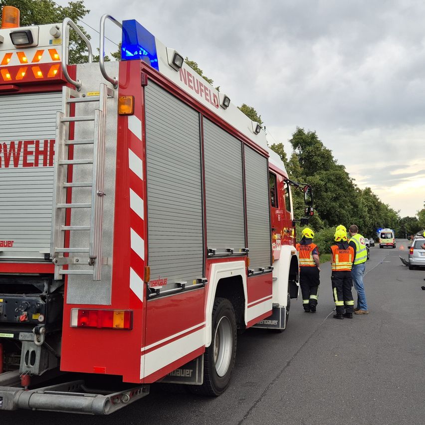 Ein roter Feuerwehrwagen mit 'Neufeld' steht mit eingeschaltetem Licht am Straßenrand. Rettungskräfte stehen in der Nähe.