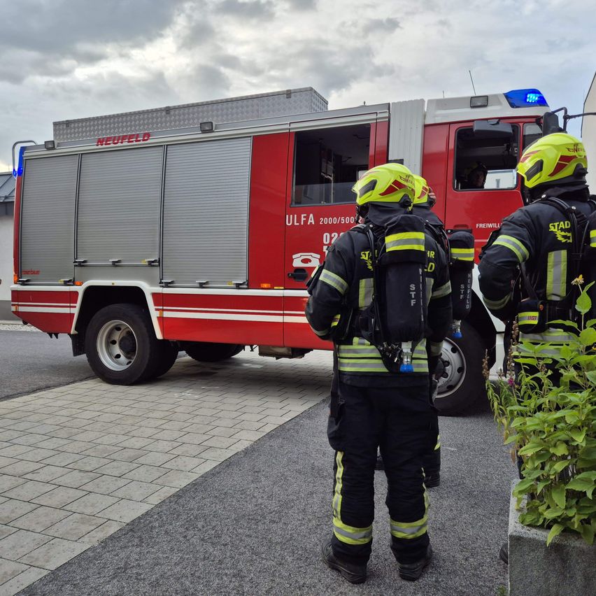 Drei Feuerwehrleute in gelben Helmen stehen neben einem roten Neufeld-Feuerwehrauto mit der Nummer 02. Sie stehen vor einem Gebäude mit Pflanzen.