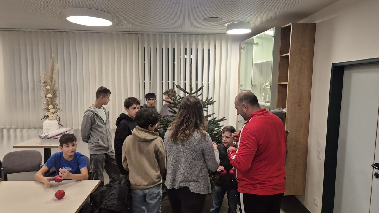 Eine Gruppe von Menschen versammelt sich um einen Weihnachtsbaum und schmückt ihn mit Ornamenten. Ein Mann in einem roten Jacket hilft einem jungen Jungen. Der Raum hat weiße Jalousien und einen Holzschrank.