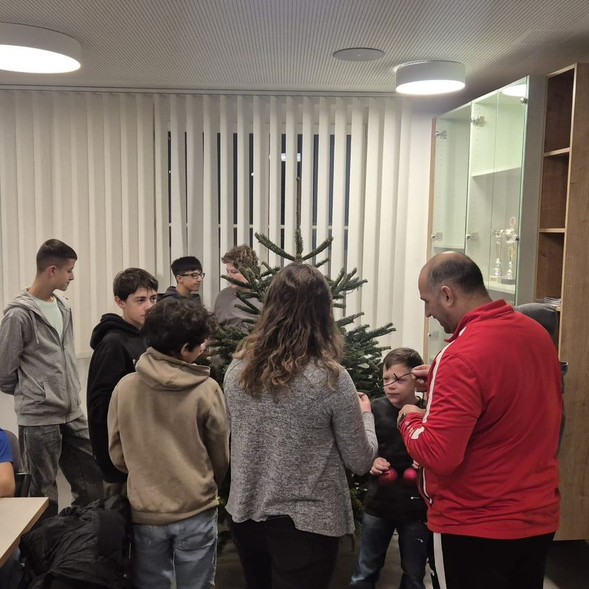 Eine Gruppe von Menschen versammelt sich um einen Weihnachtsbaum und schmückt ihn mit Ornamenten. Ein Mann in einem roten Jacket hilft einem jungen Jungen. Der Raum hat weiße Jalousien und einen Holzschrank.