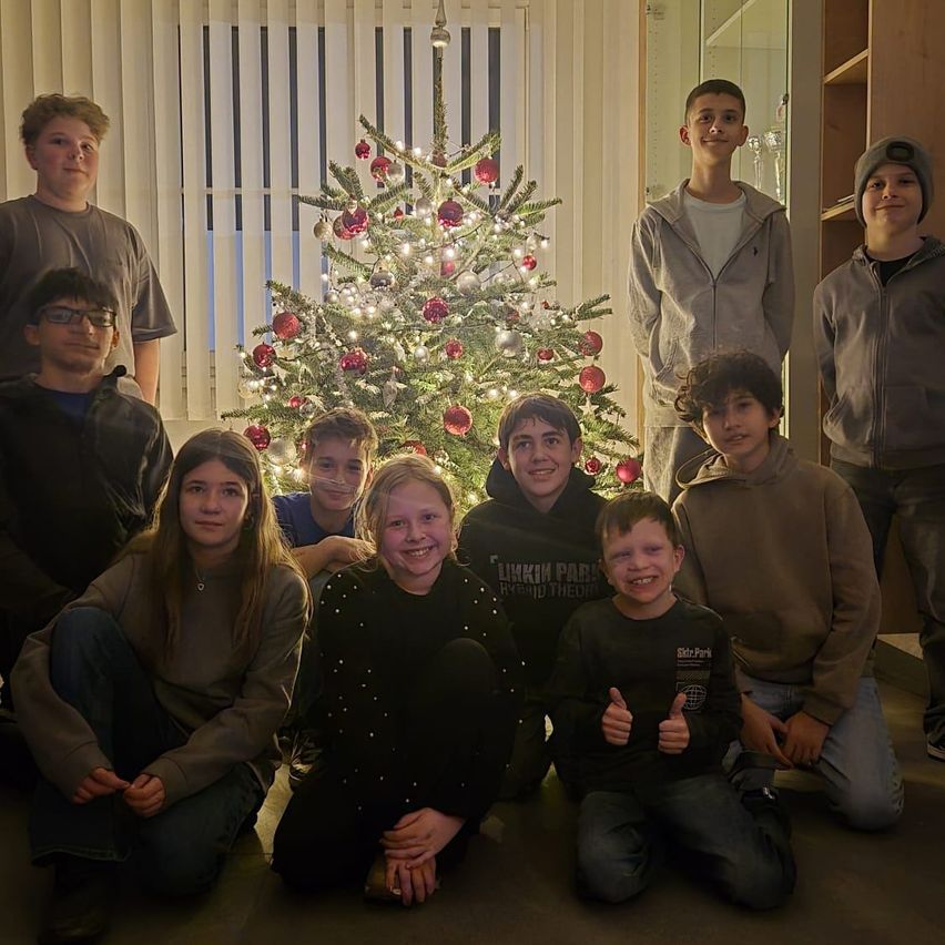 Eine Gruppe junger Menschen posiert vor einem geschmückten Weihnachtsbaum für ein Foto. Sie sitzen und stehen um den Baum herum und lächeln für die Kamera.