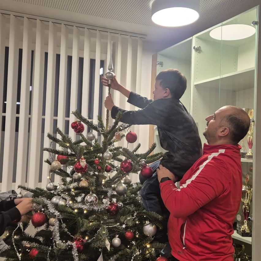 Ein Mann und ein Junge schmücken einen Weihnachtsbaum. Der Junge platziert eine silberne Kugel am Baum, während der Mann ihn von unten unterstützt.