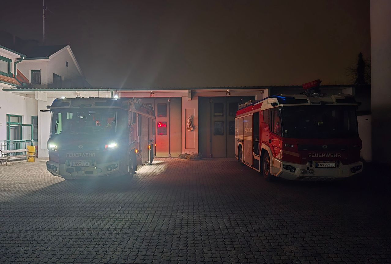 Zwei Feuerwehrfahrzeuge sind nachts vor einem Gebäude geparkt. Das vordere Fahrzeug hat seine Scheinwerfer eingeschaltet, während das hintere Fahrzeug seine Einsatzleuchten aktiviert hat.