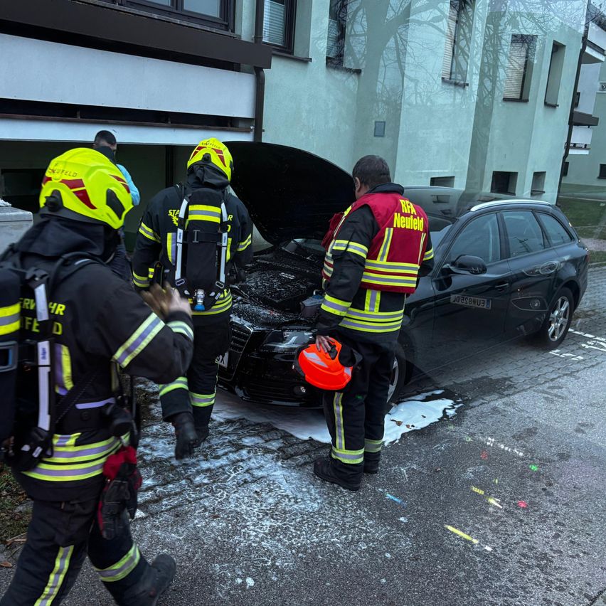 Feuerwehrleute stehen um ein schwarzes Auto mit offenem Motorhaube, am Straßenrand, einer hält einen Helm.