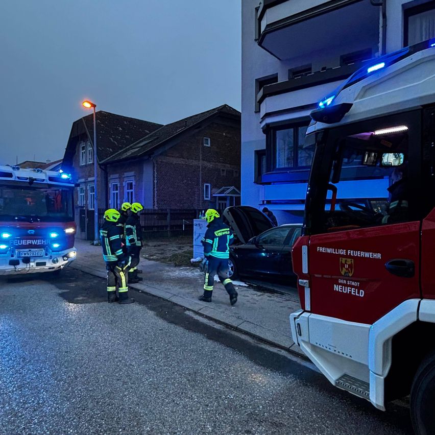 Feuerwehrleute in reflektierenden Westen stehen auf dem Bürgersteig in der Nähe eines geparkten Autos. Ein Feuerwehrwagen ist auf der Straße, seine Lichter sind eingeschaltet.